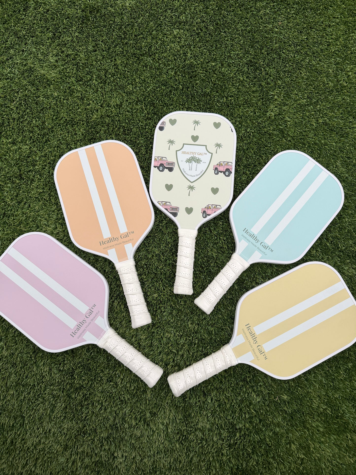 Five Healthy Gal™ Move pickleball paddles fanned out on grass showing all colorways: peach, limited-edition print, sage blue, dusty pink, and butter yellow