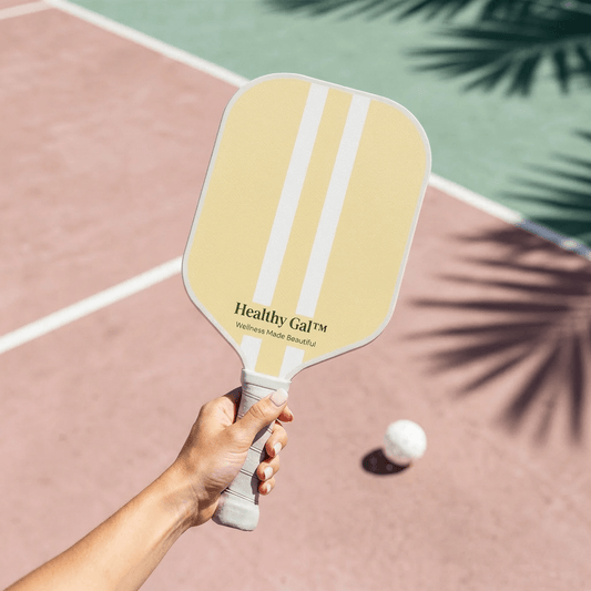 Hand holding a Healthy Gal™ Move pickleball paddle on a pink clay court with a pickleball in mid-air and palm leaf shadows