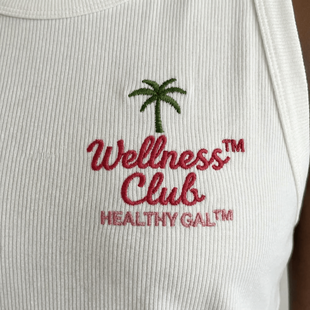 Extreme close-up of the Healthy Gal™ Wellness Club Tank embroidery — green palm tree above red script "Wellness™ Club Healthy Gal™" on white ribbed fabric