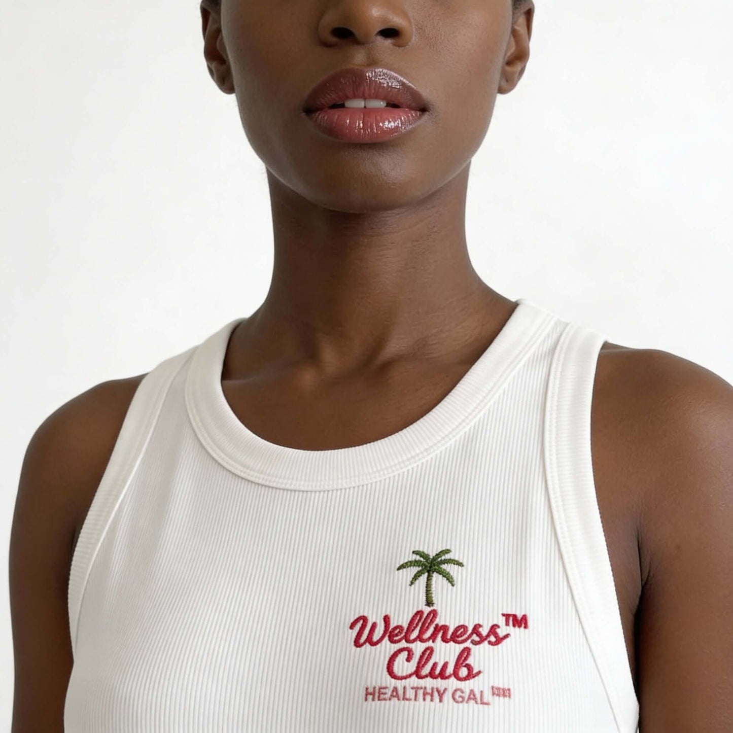 Close-up of the Healthy Gal™ Wellness Club Tank chest embroidery showing the palm tree and "Wellness Club Healthy Gal™" varsity logo on white ribbed fabric