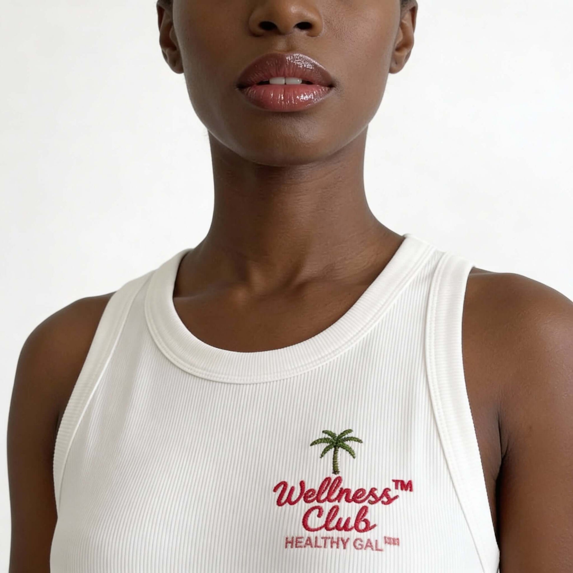 Close-up of the Healthy Gal™ Wellness Club Tank chest embroidery showing the palm tree and "Wellness Club Healthy Gal™" varsity logo on white ribbed fabric